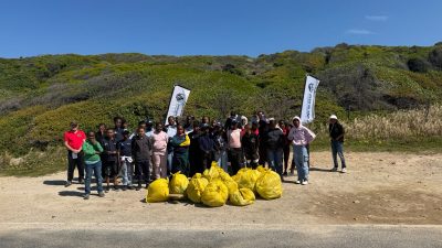 A large group of volunteers, most of them schoolchildren in warm clothing, stand on a gravel verge behind a pile of full yellow waste bags, with two banners and green coastal fynbos on the slope behind them.
