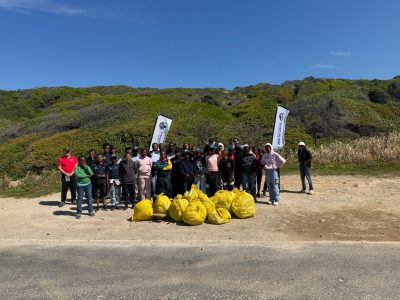 A large group of volunteers, most of them schoolchildren in warm clothing, stand on a gravel verge behind a pile of full yellow waste bags, with two banners and green coastal fynbos on the slope behind them.