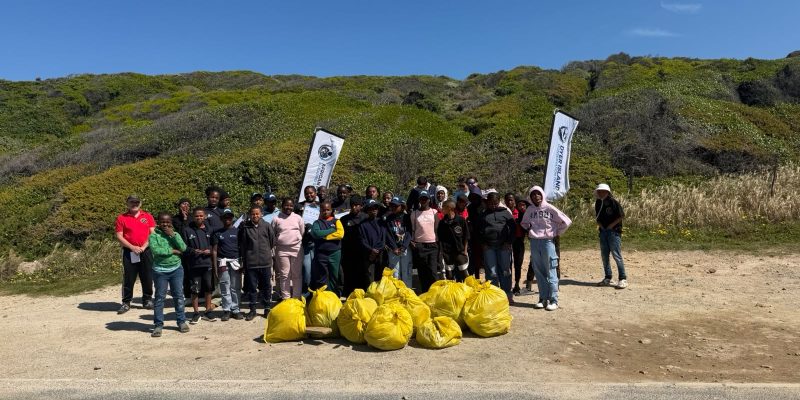 A large group of volunteers, most of them schoolchildren in warm clothing, stand on a gravel verge behind a pile of full yellow waste bags, with two banners and green coastal fynbos on the slope behind them.