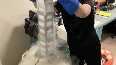 A researcher in heavy blue cryogenic gloves lifts a tall rack of labeled sample canes out of a liquid nitrogen storage tank in a laboratory, with white vapor pouring down over the tank and across the floor.