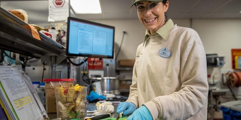 A Disney cast member slicing fruit beside a screen showing an animal diet plan