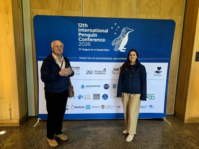 A man and a woman stand either side of a large blue banner reading 12th International Penguin Conference 2026, 31 August to 4 September.