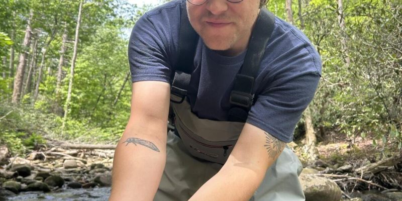 A person in waders standing in a forest creek holds a small eastern hellbender salamander curled in one cupped hand.
