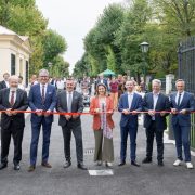 Seven people cutting a red ribbon across an open iron gate leading onto a tree-lined avenue