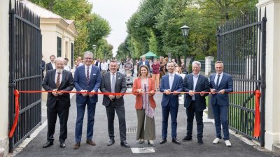 Seven people cutting a red ribbon across an open iron gate leading onto a tree-lined avenue