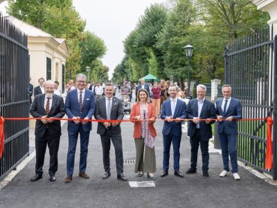 Seven people cutting a red ribbon across an open iron gate leading onto a tree-lined avenue