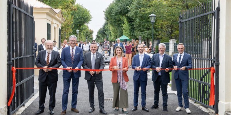 Seven people cutting a red ribbon across an open iron gate leading onto a tree-lined avenue