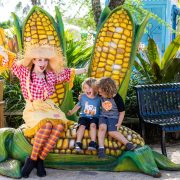 A performer in a scarecrow costume sits on a giant corn sculpture with two laughing children at ZooTampa
