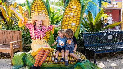 A performer in a scarecrow costume sits on a giant corn sculpture with two laughing children at ZooTampa