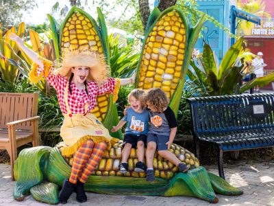 A performer in a scarecrow costume sits on a giant corn sculpture with two laughing children at ZooTampa