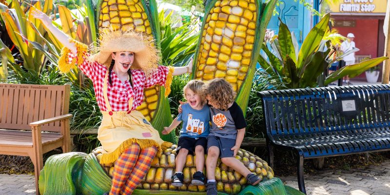 A performer in a scarecrow costume sits on a giant corn sculpture with two laughing children at ZooTampa