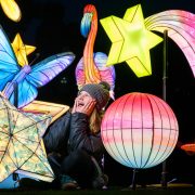 A child in a woolly hat sitting among lit star, planet, butterfly and flamingo lanterns at night
