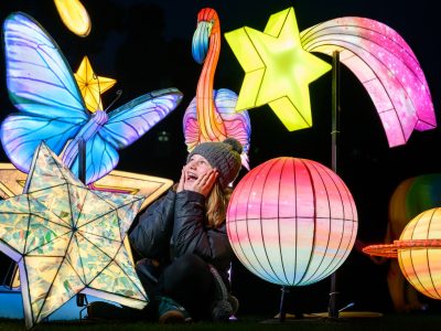 A child in a woolly hat sitting among lit star, planet, butterfly and flamingo lanterns at night
