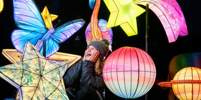 A child in a woolly hat sitting among lit star, planet, butterfly and flamingo lanterns at night