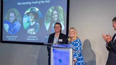 Two speakers stand at a lectern bearing the Loggerhead Marinelife Center logo, with a large screen behind them reading 2026 Go Blue Awards Finalists and Blue Hatchling Youth of the Year above three circular portraits of the youth finalists, while a man at the right applauds.