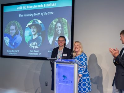 Two speakers stand at a lectern bearing the Loggerhead Marinelife Center logo, with a large screen behind them reading 2026 Go Blue Awards Finalists and Blue Hatchling Youth of the Year above three circular portraits of the youth finalists, while a man at the right applauds.