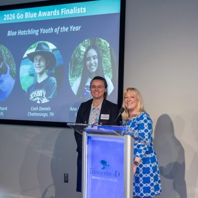 Two speakers stand at a lectern bearing the Loggerhead Marinelife Center logo, with a large screen behind them reading 2026 Go Blue Awards Finalists and Blue Hatchling Youth of the Year above three circular portraits of the youth finalists, while a man at the right applauds.