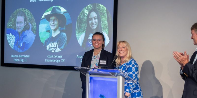 Two speakers stand at a lectern bearing the Loggerhead Marinelife Center logo, with a large screen behind them reading 2026 Go Blue Awards Finalists and Blue Hatchling Youth of the Year above three circular portraits of the youth finalists, while a man at the right applauds.