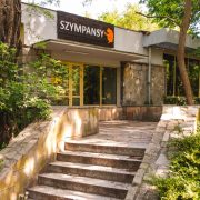 Stone steps lead up to a low modernist zoo building with a dark sign reading SZYMPANSY above its glass entrance, framed by trees.