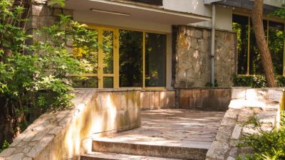 Stone steps lead up to a low modernist zoo building with a dark sign reading SZYMPANSY above its glass entrance, framed by trees.