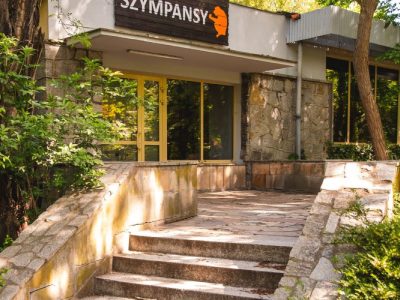Stone steps lead up to a low modernist zoo building with a dark sign reading SZYMPANSY above its glass entrance, framed by trees.