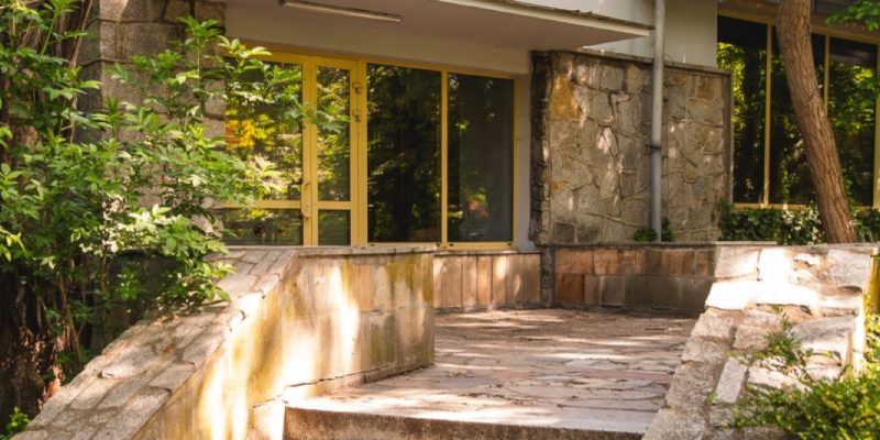 Stone steps lead up to a low modernist zoo building with a dark sign reading SZYMPANSY above its glass entrance, framed by trees.