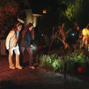 Two children in Halloween headbands walk a gravel path at night with a flashlight, past autumn planting lit from below, dried cornstalks and a carved pumpkin, with a pale wooden house behind them.