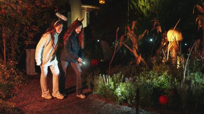 Two children in Halloween headbands walk a gravel path at night with a flashlight, past autumn planting lit from below, dried cornstalks and a carved pumpkin, with a pale wooden house behind them.