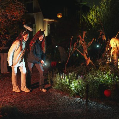 Two children in Halloween headbands walk a gravel path at night with a flashlight, past autumn planting lit from below, dried cornstalks and a carved pumpkin, with a pale wooden house behind them.