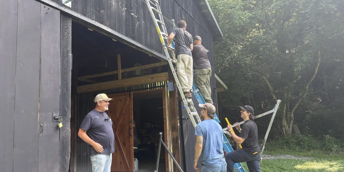 Five men mounting solar rails on the side of a dark wooden barn, two on ladders and three watching from the ground