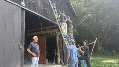 Five men mounting solar rails on the side of a dark wooden barn, two on ladders and three watching from the ground