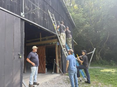 Five men mounting solar rails on the side of a dark wooden barn, two on ladders and three watching from the ground
