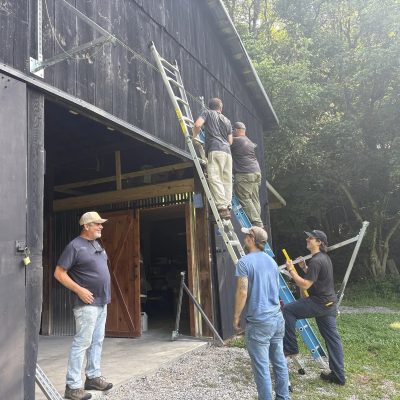 Five men mounting solar rails on the side of a dark wooden barn, two on ladders and three watching from the ground