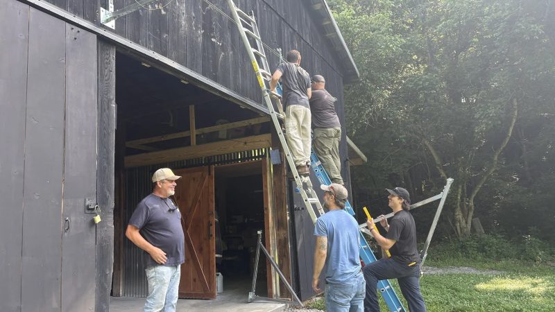 Five men mounting solar rails on the side of a dark wooden barn, two on ladders and three watching from the ground