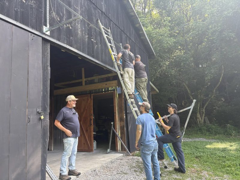 Five men mounting solar rails on the side of a dark wooden barn, two on ladders and three watching from the ground
