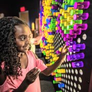 A child presses a purple pin into a wall-sized grid filled with glowing coloured light pins in a darkened gallery.