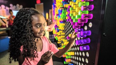 A child presses a purple pin into a wall-sized grid filled with glowing coloured light pins in a darkened gallery.