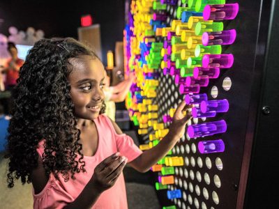 A child presses a purple pin into a wall-sized grid filled with glowing coloured light pins in a darkened gallery.