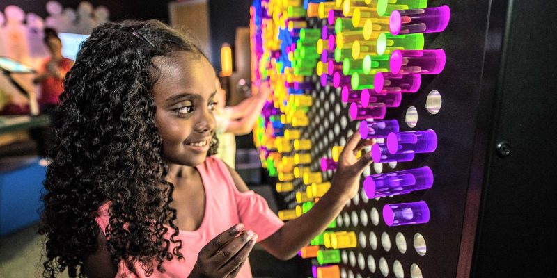 A child presses a purple pin into a wall-sized grid filled with glowing coloured light pins in a darkened gallery.