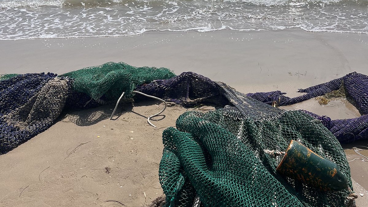 Large tangled masses of discarded green and dark blue fishing net, one still attached to a barnacle-crusted float, lie washed up on wet sand at the waterline