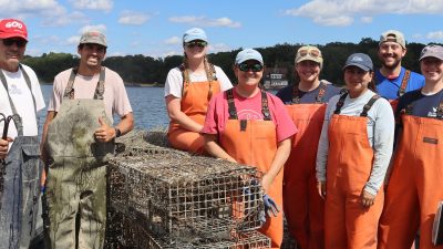 Nine people in orange and tan waders stand smiling on the deck of a boat on Long Island Sound behind a stack of encrusted lobster traps recovered from the seafloor, with a wooded shoreline behind them