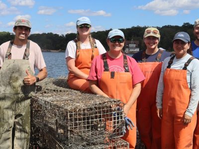 Nine people in orange and tan waders stand smiling on the deck of a boat on Long Island Sound behind a stack of encrusted lobster traps recovered from the seafloor, with a wooded shoreline behind them