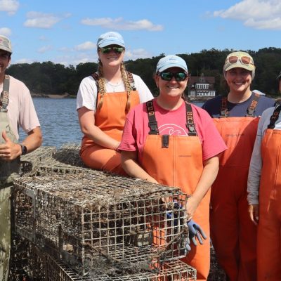Nine people in orange and tan waders stand smiling on the deck of a boat on Long Island Sound behind a stack of encrusted lobster traps recovered from the seafloor, with a wooded shoreline behind them