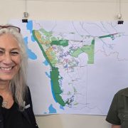 Two women stand smiling on either side of a large printed map of Namibia pinned to a wall, the map marked with green protected areas and conservancy boundaries; the woman on the left wears a black Cheetah Conservation Fund shirt and the woman on the right a green WWF shirt.