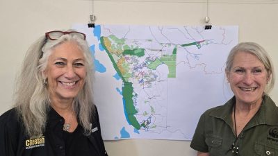Two women stand smiling on either side of a large printed map of Namibia pinned to a wall, the map marked with green protected areas and conservancy boundaries; the woman on the left wears a black Cheetah Conservation Fund shirt and the woman on the right a green WWF shirt.