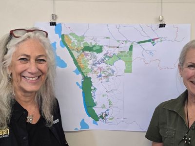 Two women stand smiling on either side of a large printed map of Namibia pinned to a wall, the map marked with green protected areas and conservancy boundaries; the woman on the left wears a black Cheetah Conservation Fund shirt and the woman on the right a green WWF shirt.