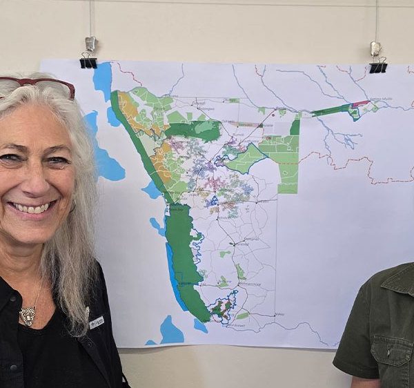 Two women stand smiling on either side of a large printed map of Namibia pinned to a wall, the map marked with green protected areas and conservancy boundaries; the woman on the left wears a black Cheetah Conservation Fund shirt and the woman on the right a green WWF shirt.