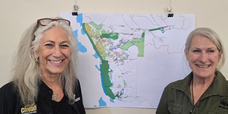 Two women stand smiling on either side of a large printed map of Namibia pinned to a wall, the map marked with green protected areas and conservancy boundaries; the woman on the left wears a black Cheetah Conservation Fund shirt and the woman on the right a green WWF shirt.