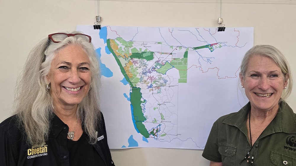 Two women stand smiling on either side of a large printed map of Namibia pinned to a wall, the map marked with green protected areas and conservancy boundaries; the woman on the left wears a black Cheetah Conservation Fund shirt and the woman on the right a green WWF shirt.