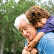 A smiling child rides piggyback on an older man's shoulders outdoors, with green trees behind them.